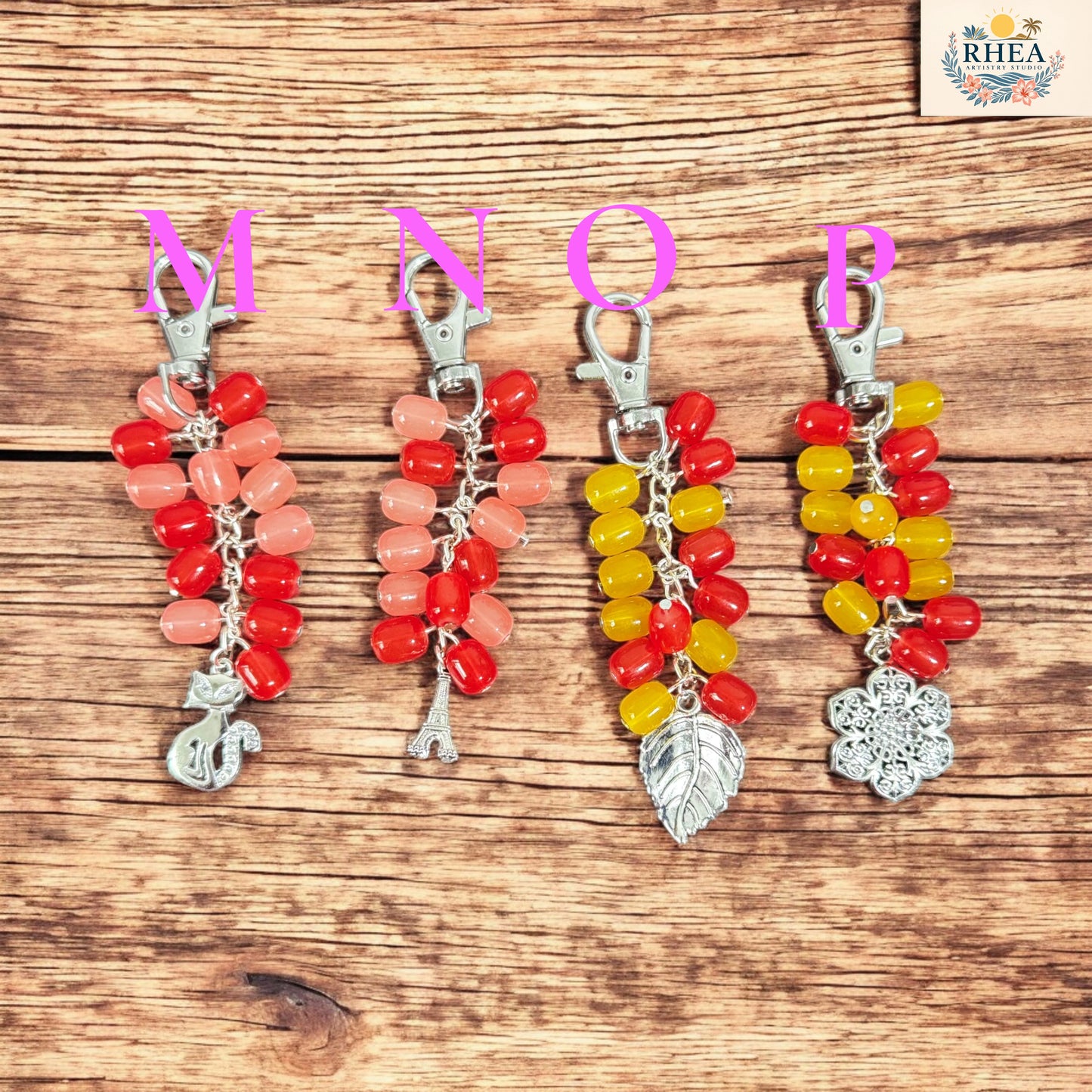 Four colorful keychains with letter charms on a wooden surface, branded 'RHEA'.