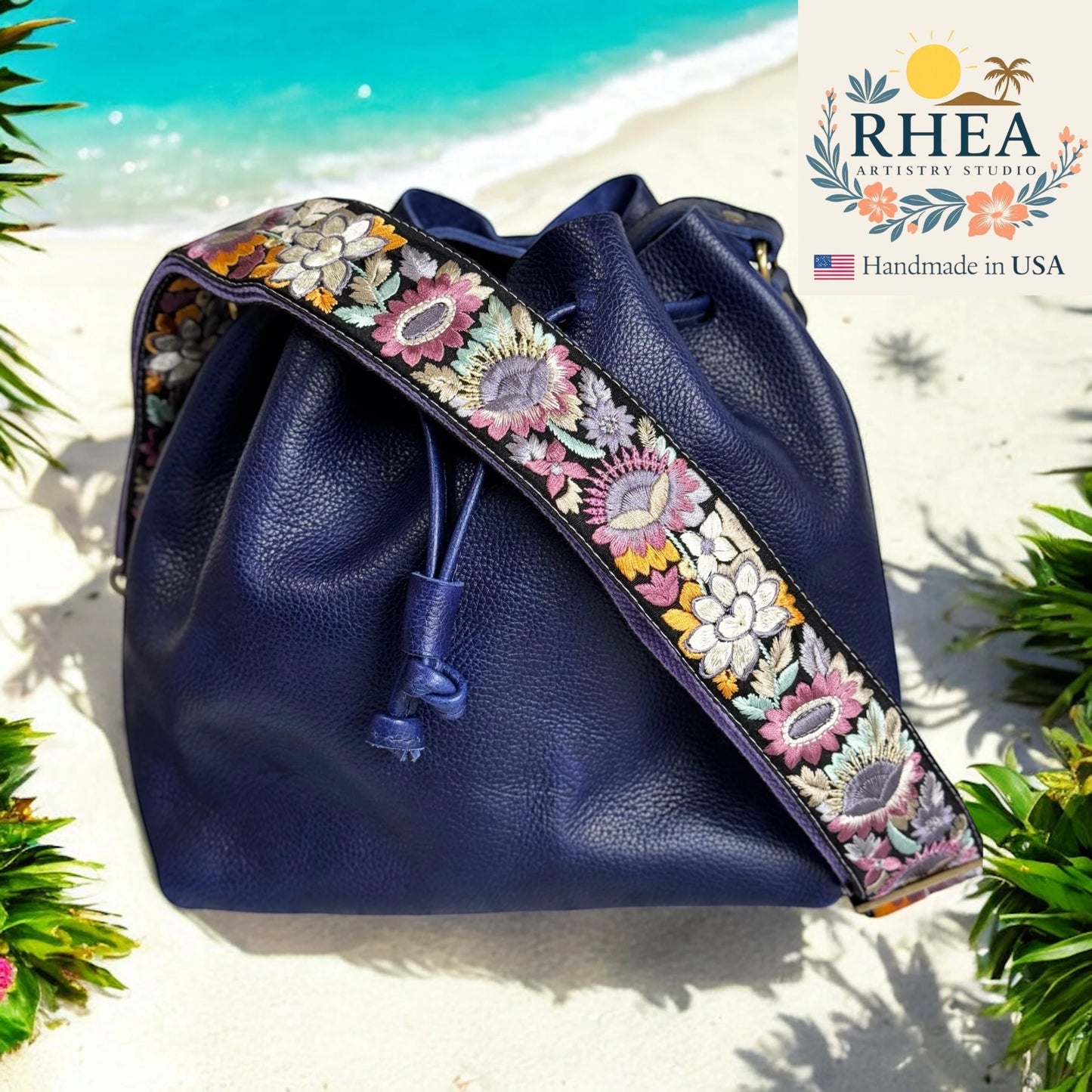Navy blue leather bag with floral embroidery purse strap on a beach background, featuring the RHEA Artistry Studio logo.