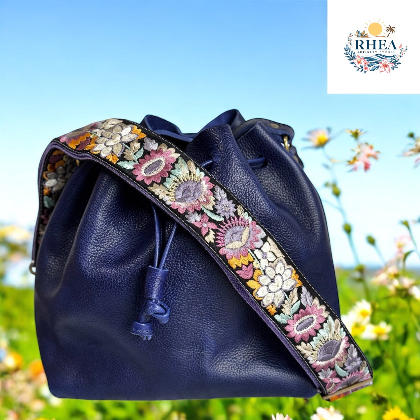 Lavender and yellow embroidered sunflower strap on black webbing, attached to navy leather bucket bag on meadow