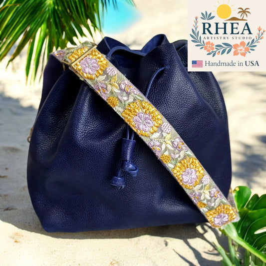 Navy blue leather bag with a decorative floral purse strap on a sandy background, featuring Rhea Artistry Studio branding.