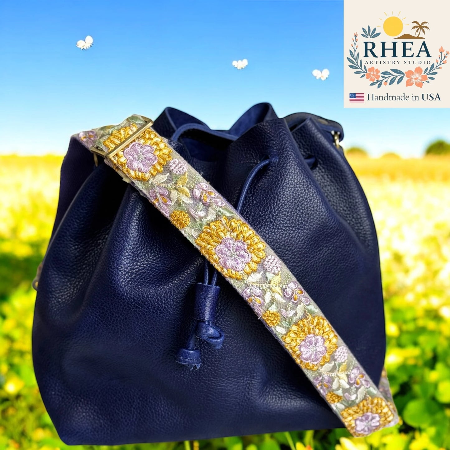 Navy blue leather bag with a floral purse strap against a blurred natural background, featuring the RHEA brand.