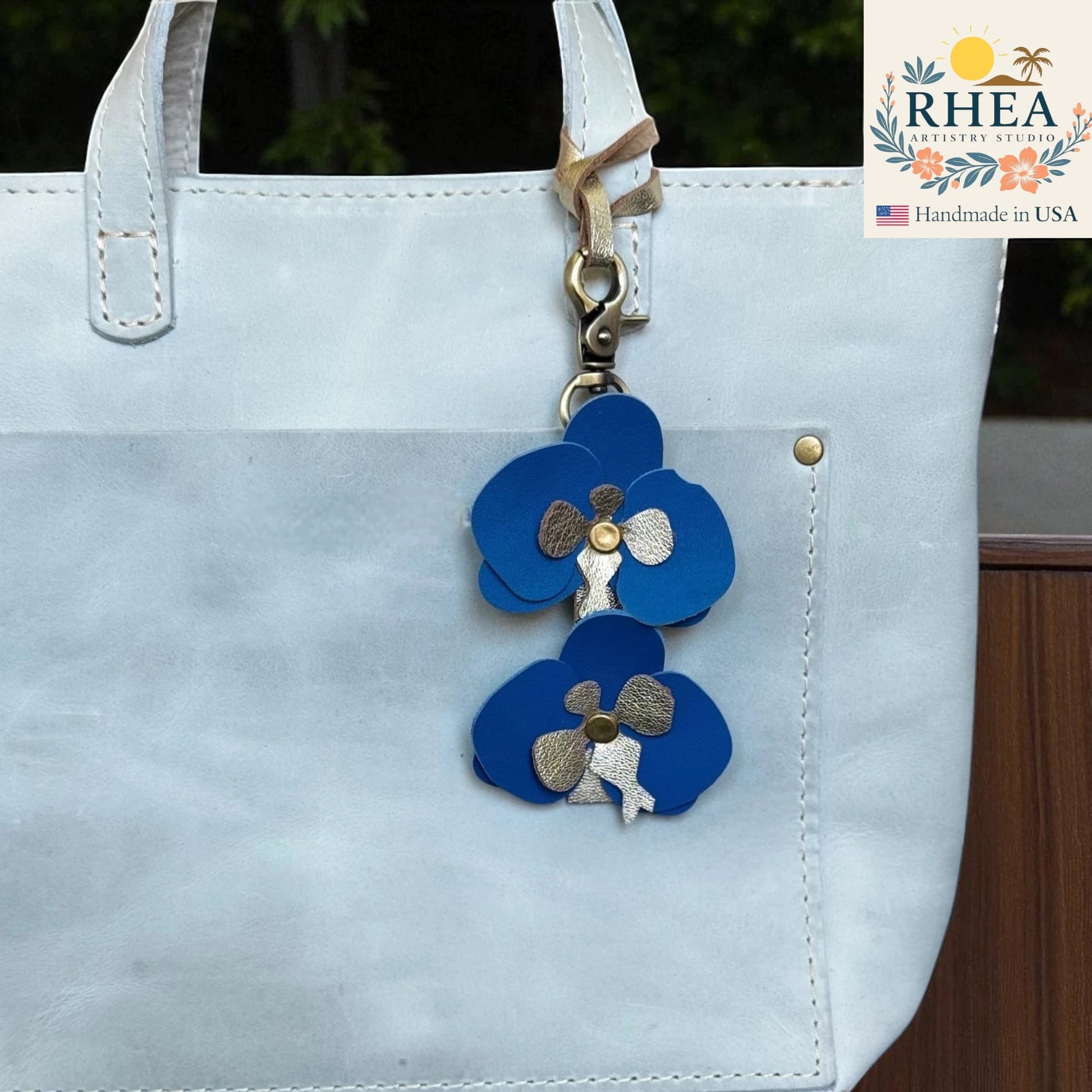 Blue flower-shaped leather bag charm attached to a white bag with RHEA branding.