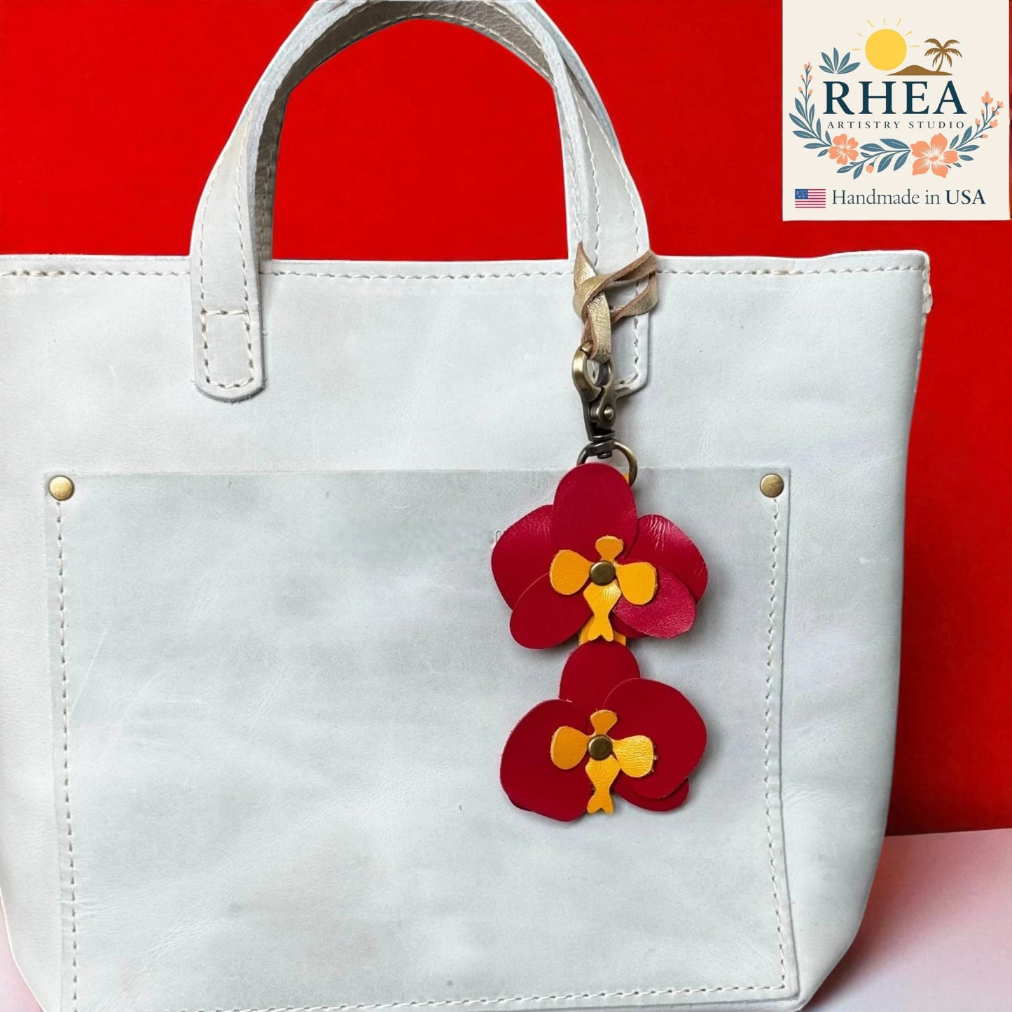 White handbag with a red and yellow floral leather purse charm on a red background, featuring the RHEA Artisan Studio logo.