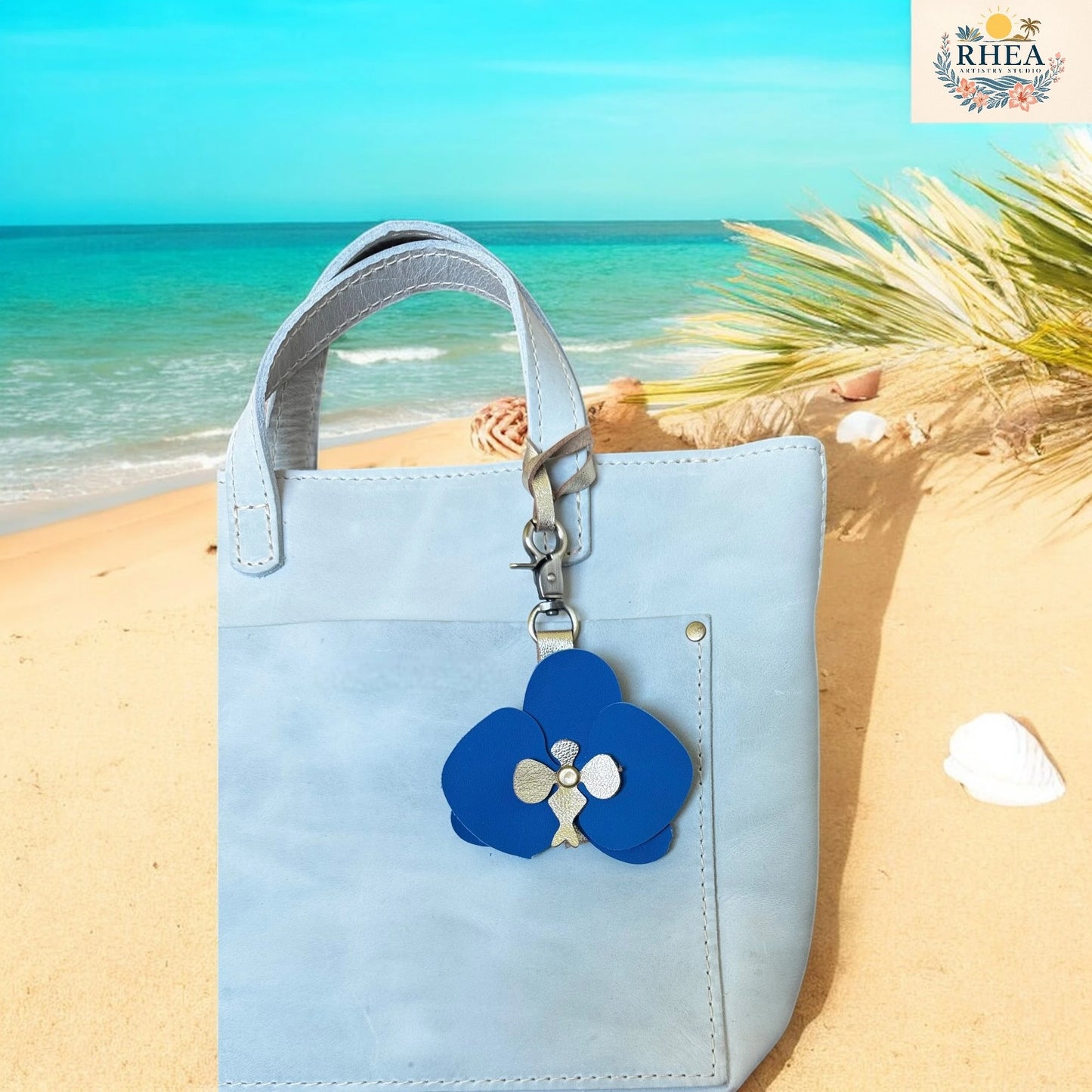 Light blue handbag with a blue flower keychain on a sandy beach with ocean view