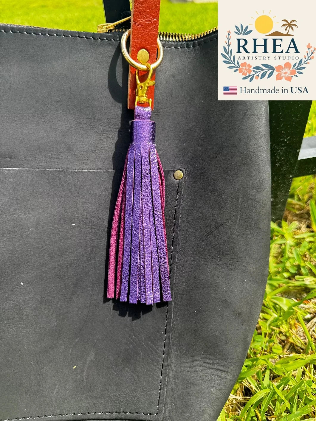Black leather bag with a purple tassel on a grassy background, featuring RHEEA branding.