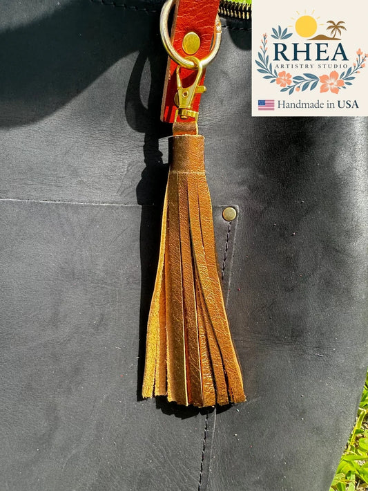 bronze leather tassel attached to a dark bag with RHEA Studio branding.