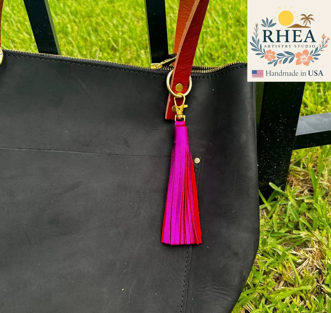 Black bag with a pink tassel on a grassy background, featuring the RHEA brand.