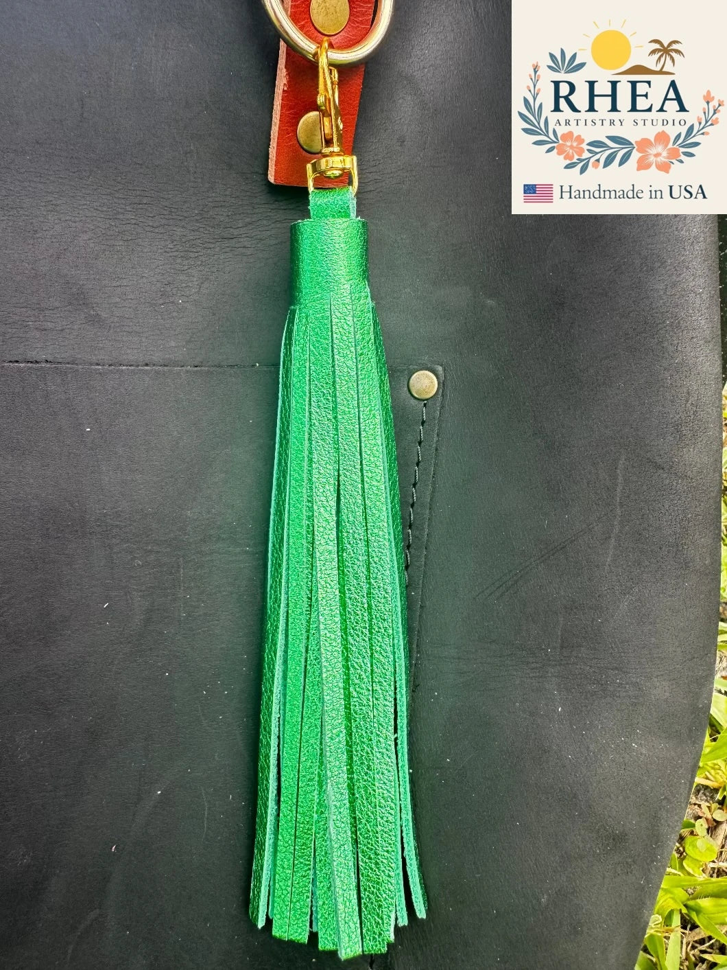 Green tassel on a black leather bag with RHEA Artisans Studio branding.