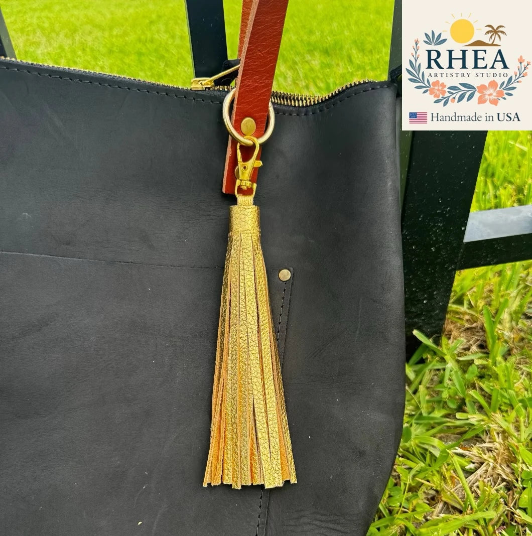 Black handbag with a gold tassel on a grassy background, featuring the RHEA brand.