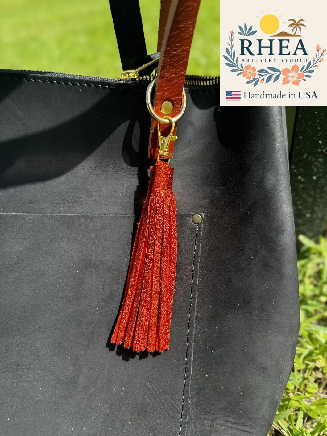 Black leather bag with a orange tassel and 'RHEA' branding in the corner.