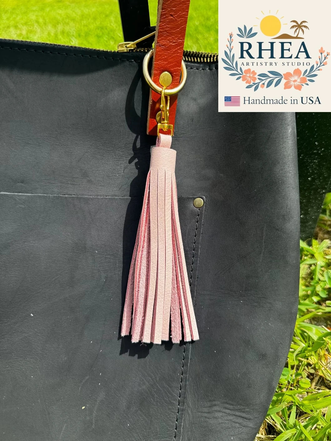 Black leather bag with a pink tassel and brown strap, featuring the RHEA Artistry Studios logo.