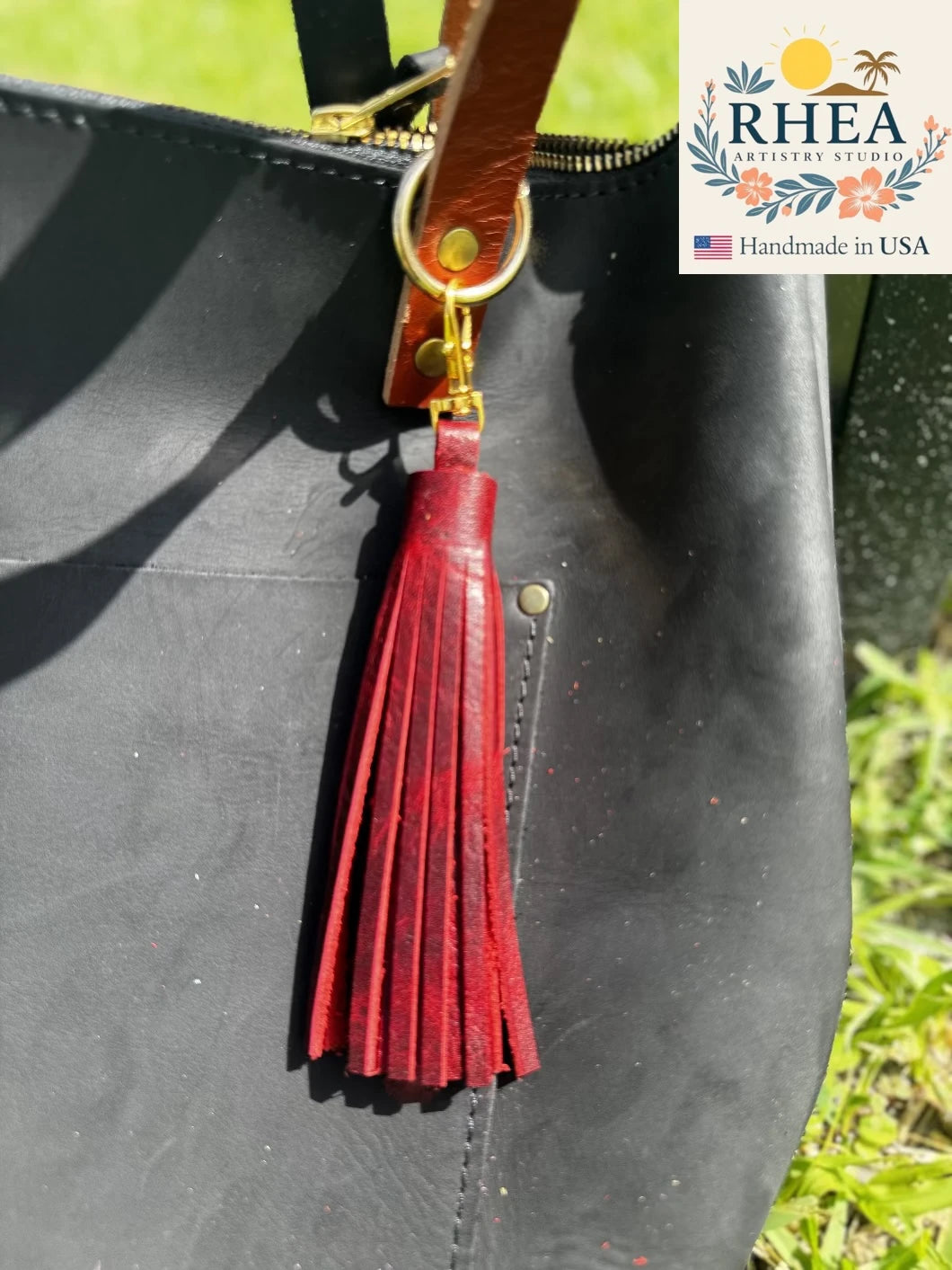 Black leather bag with a sangria tassel and gold zipper pull, featuring the RHEA brand logo.