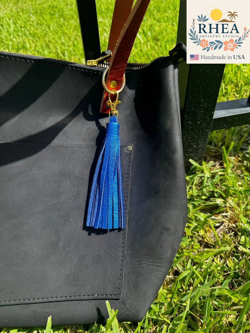 Black handbag with a blue tassel on grass, RHEA brand visible