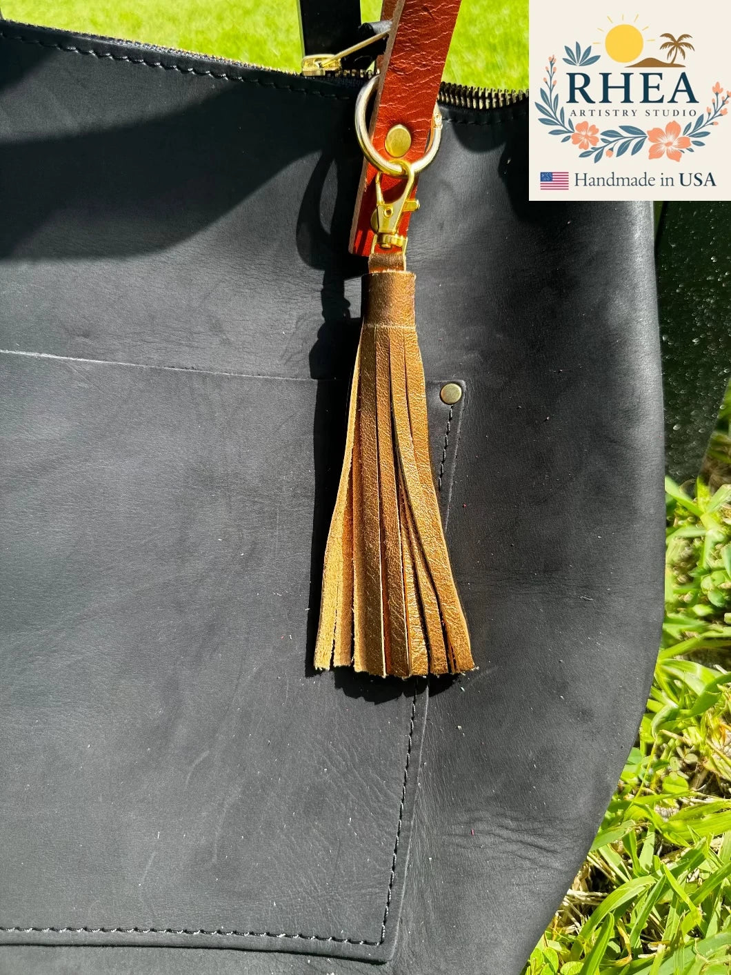 Black leather bag with a bronze leather tassel and RHEA branding.