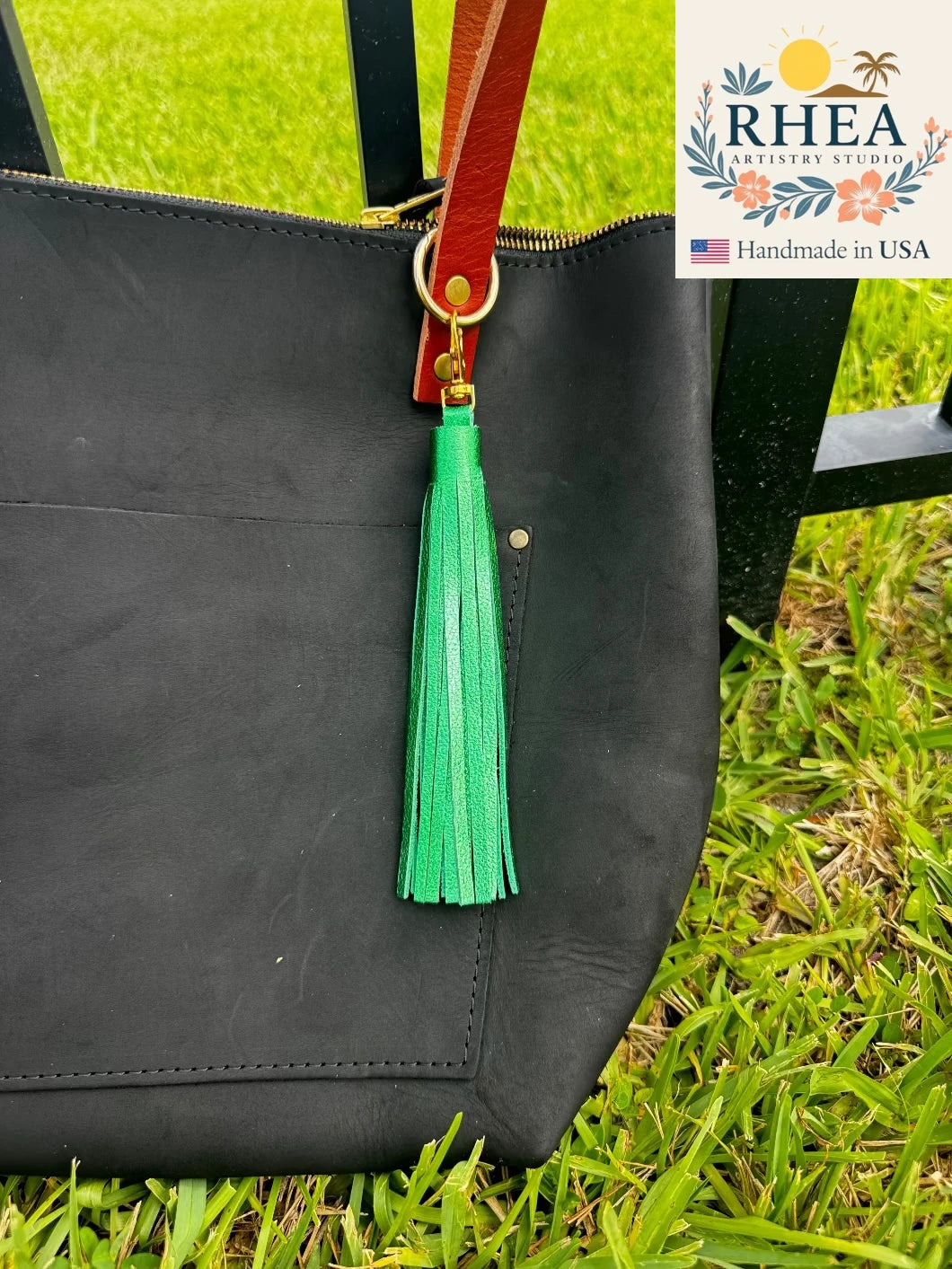 Black leather tote bag with a emerald green tassel on grass, featuring RHEA branding.
