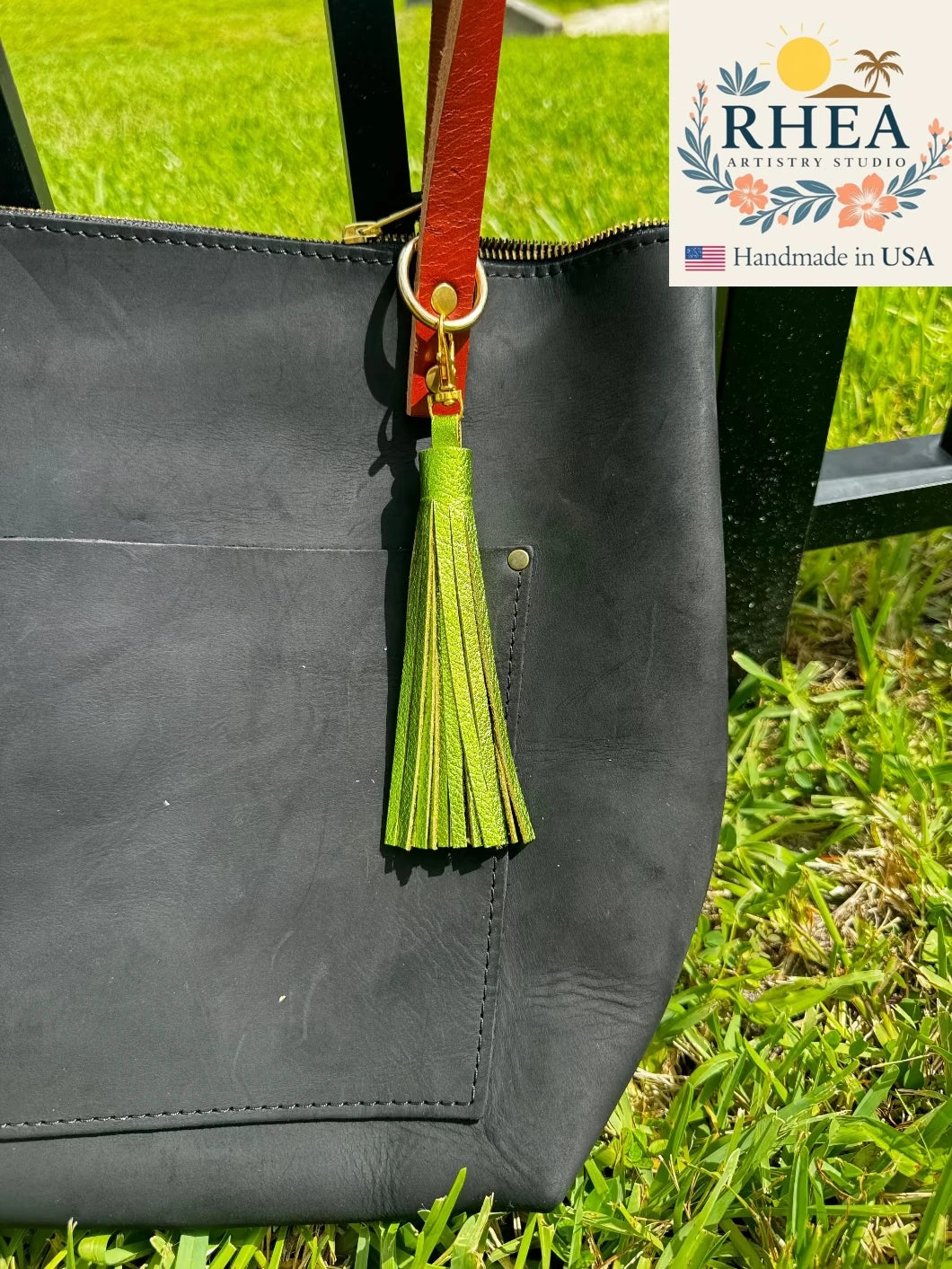Black bag with a fern green tassel on grass, featuring RHEA Artisan Studio branding.