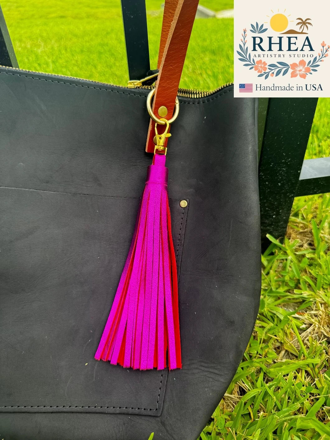 Black handbag with a pink tassel on a grassy background, featuring RHEA Artisan Studio branding.