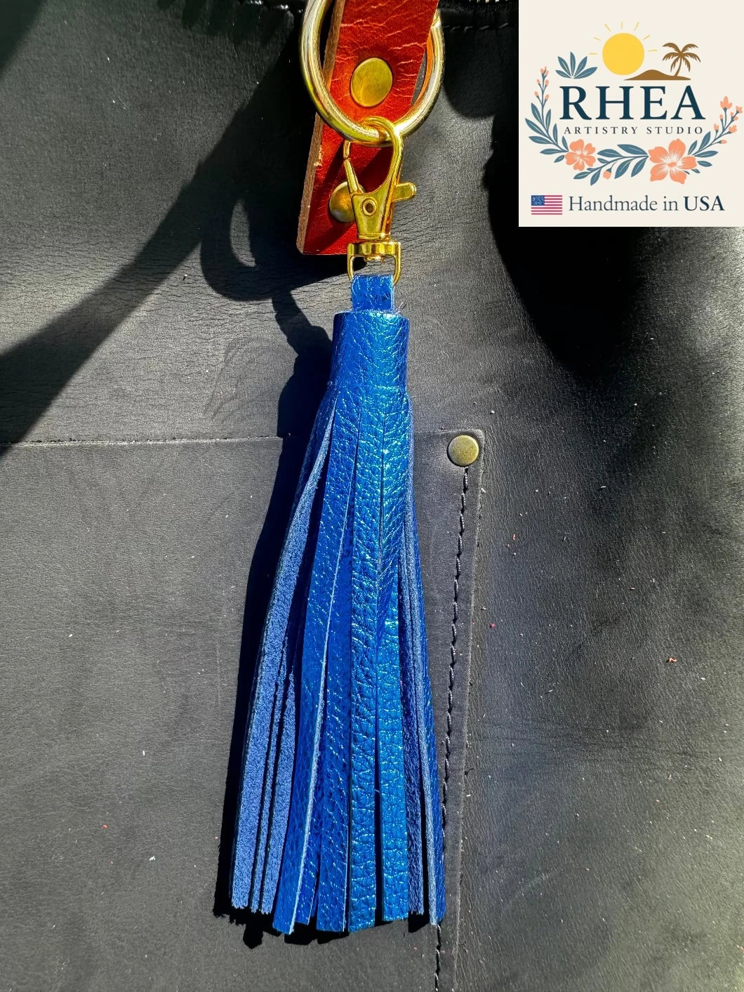 Blue leather tassel keychain on a dark surface with RHEEA branding.
