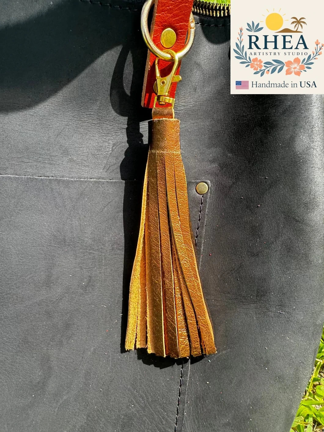 bronze leather tassel keychain attached to a black leather bag with RHEEA Artisan Studio branding.