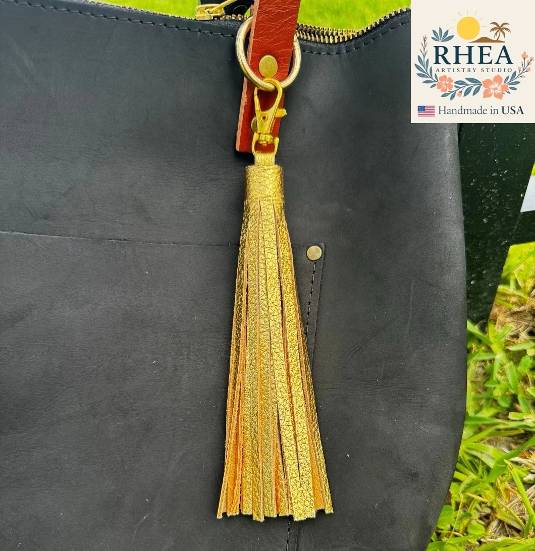 Black leather bag with a gold tassel on a grassy background, featuring the RHEA Artistry Studio logo.