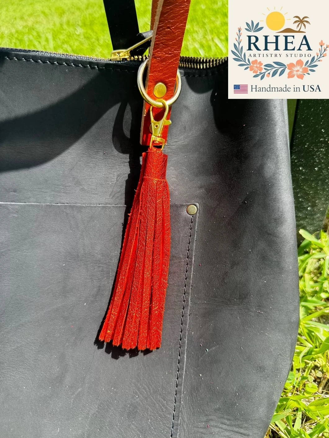Black leather bag with a orange tassel on a grassy background, featuring the RHEA Artistry Studio logo.