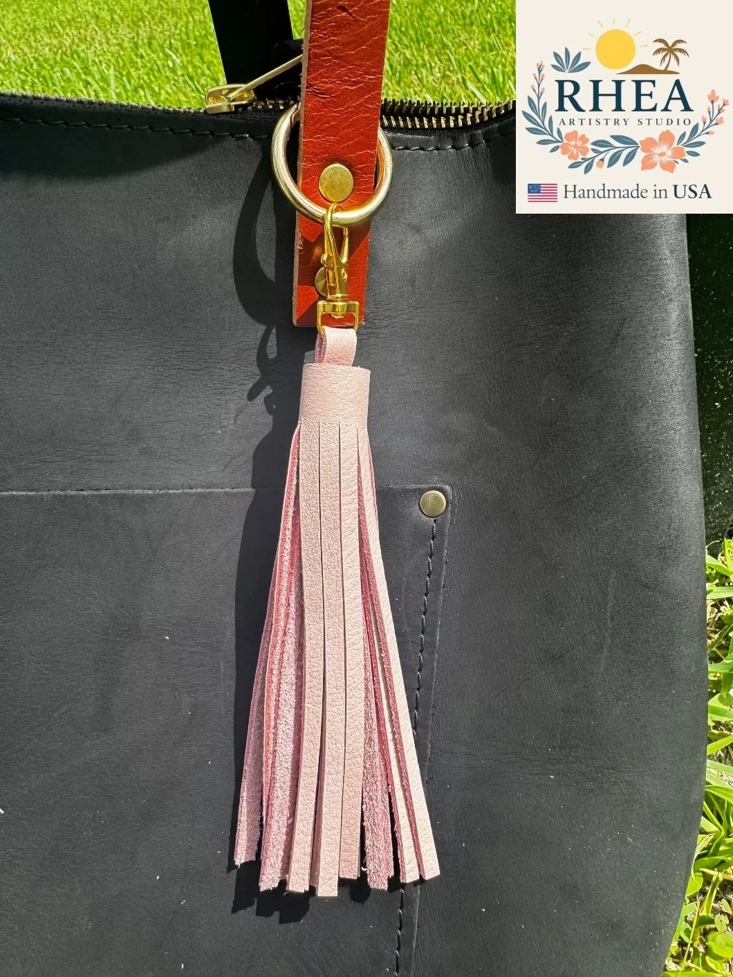 Black leather bag with a pink tassel and red leather strap, featuring the RHEA Artisan Studio logo.