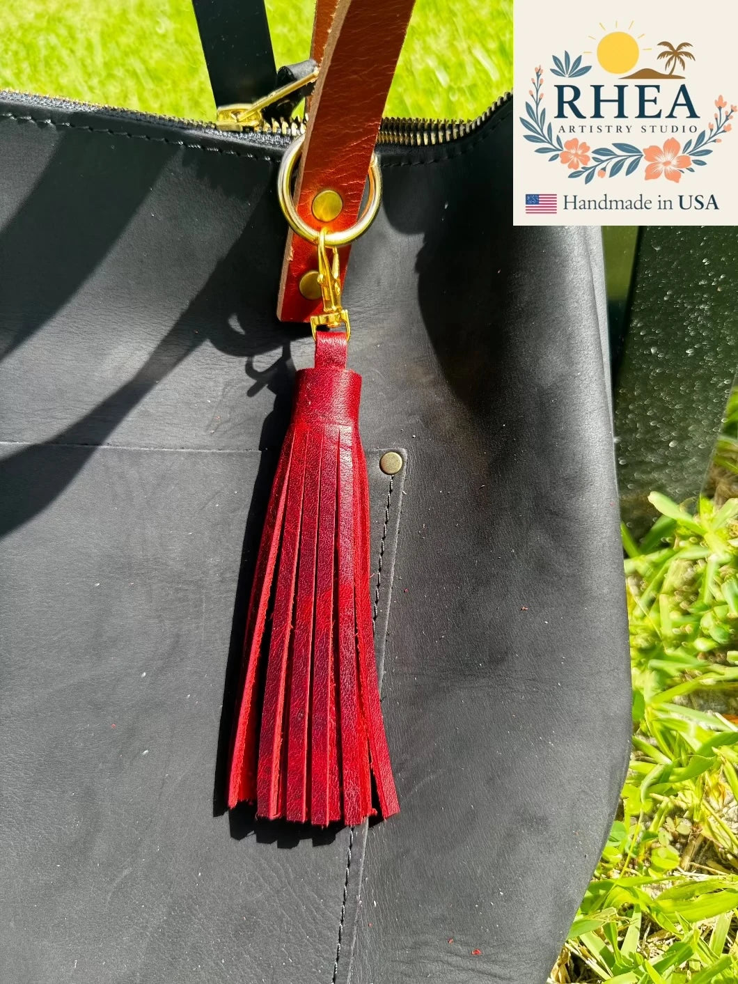 Black bag with a sangria tassel and gold zipper pull on grass, featuring RHEEA branding.