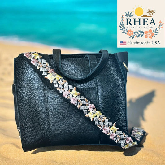 Black handbag with floral strap on a beach, featuring RHEA Accessories Studio logo.