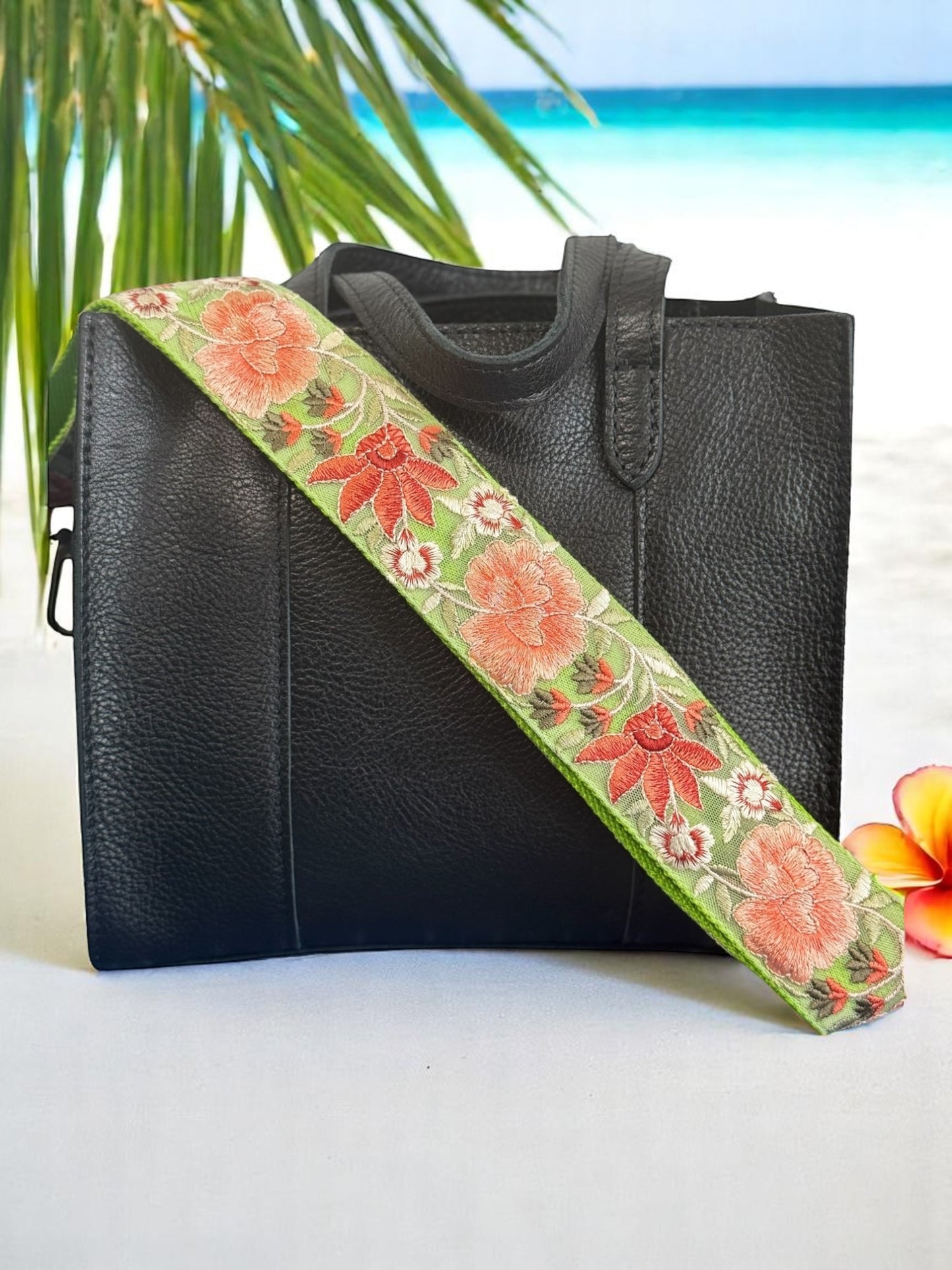 A black purse with a pink and green floral embroidered strap placed on a surface with a field of flowers in the background.