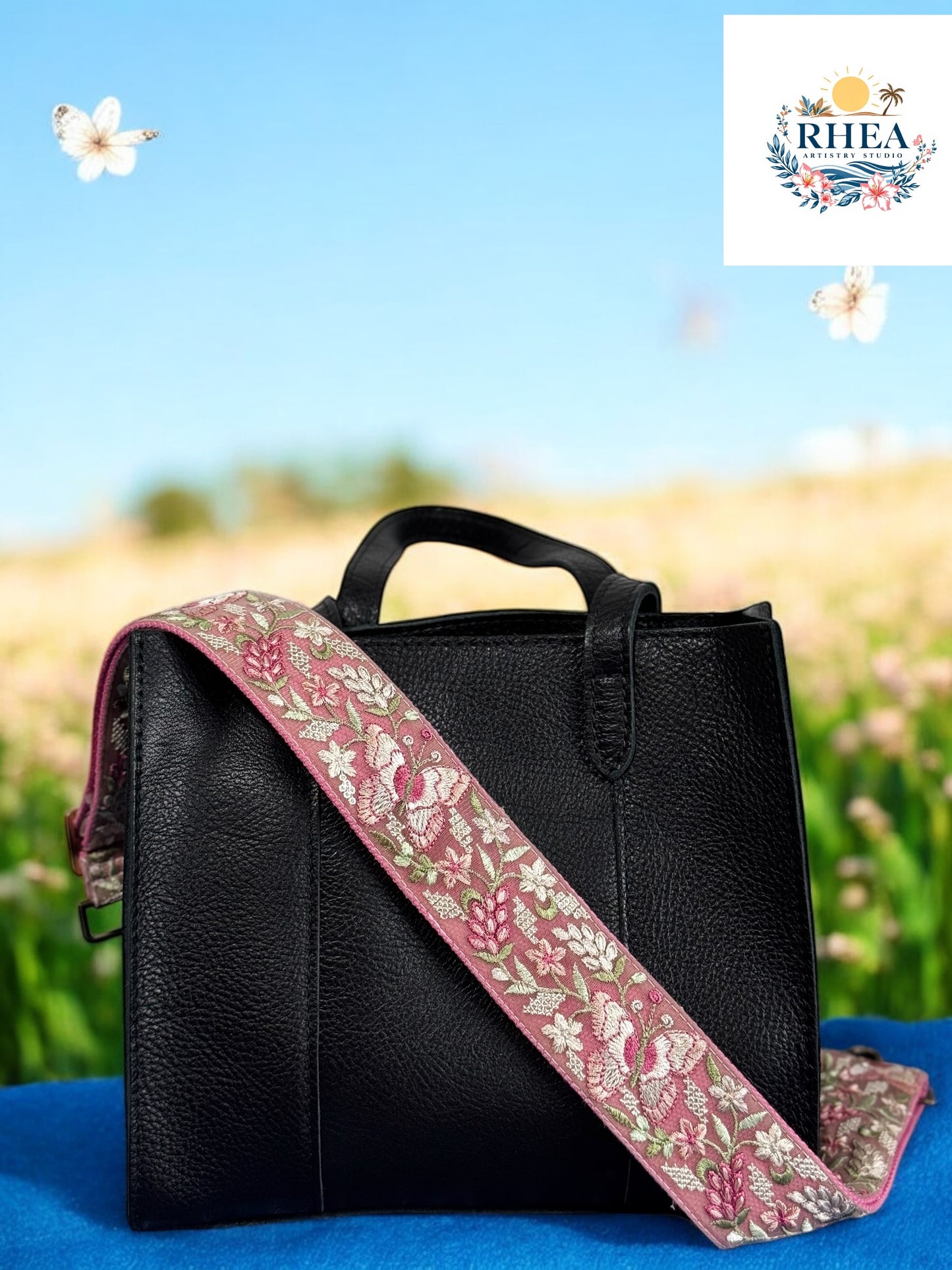 A black purse with a pink and green floral embroidered strap placed on a blue surface with flowers in the background.