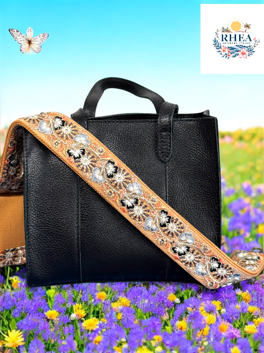 A black purse with a pink and green floral embroidered strap placed on a blue surface with a bright sky and greenery in the background.