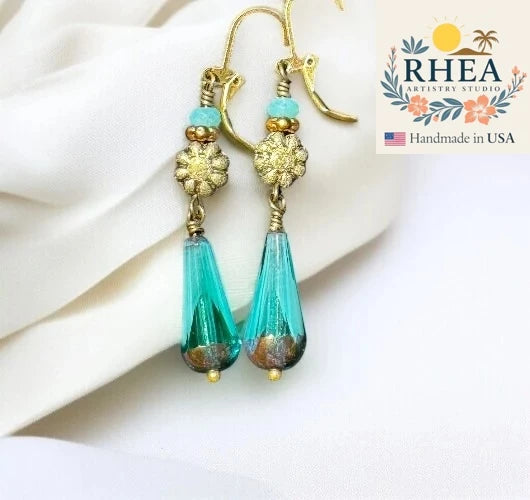 Turquoise teardrop earrings with gold accents on a white fabric background, featuring the Rhea Artistry Studio logo.