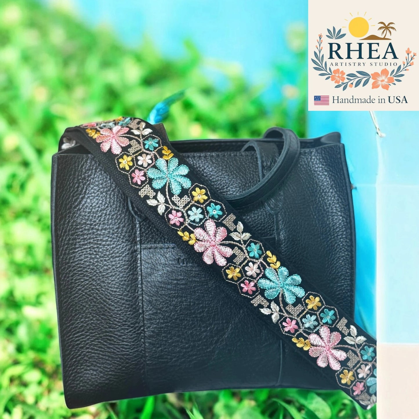 Black leather handbag with a colorful floral strap against a green grass background, featuring the RHEA Artistry Studio logo.