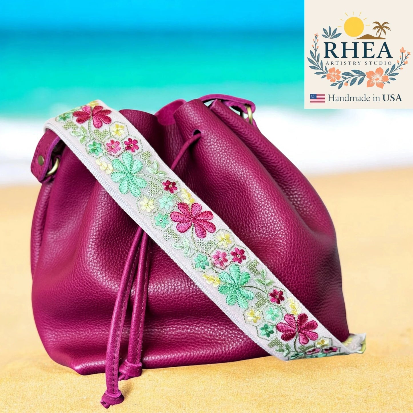 Pink leather bag with a floral strap on a beach background, featuring the RHEA Artistry Studio logo.