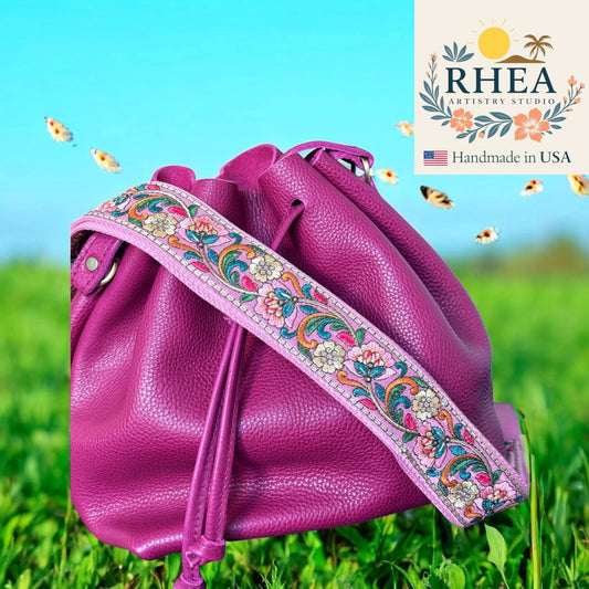 Pink leather bag with a colorful strap on grass, RHEA Artistry Studio logo visible
