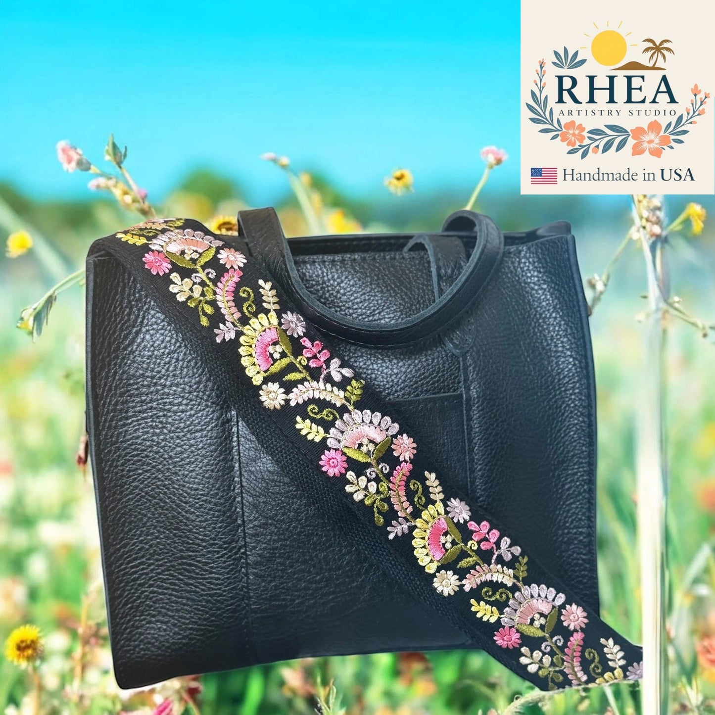 Black leather handbag with floral embroidery against a natural background, featuring the RHEA Artistry Studio logo.