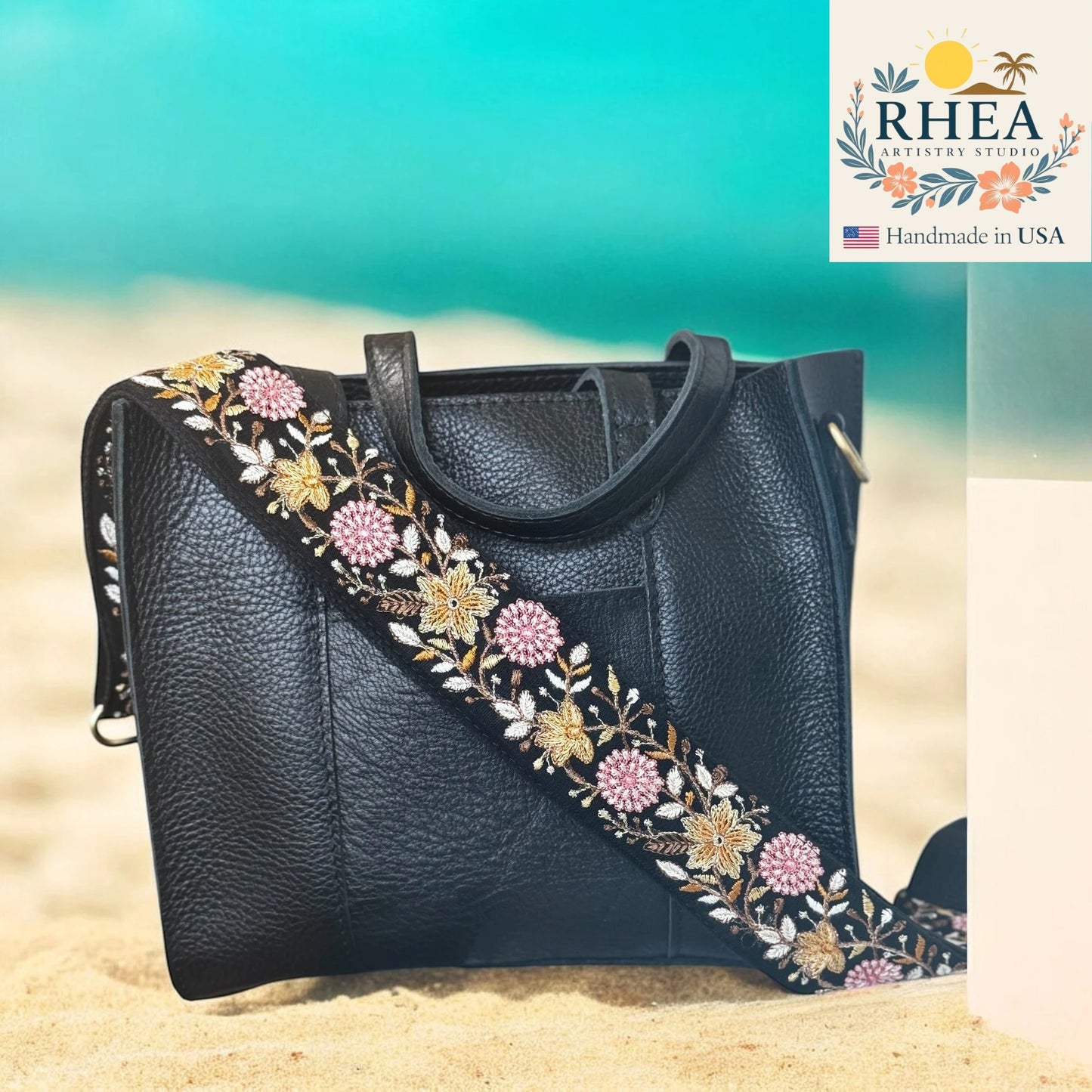 Black handbag with floral embroidery on a beach background, featuring the RHEA brand.