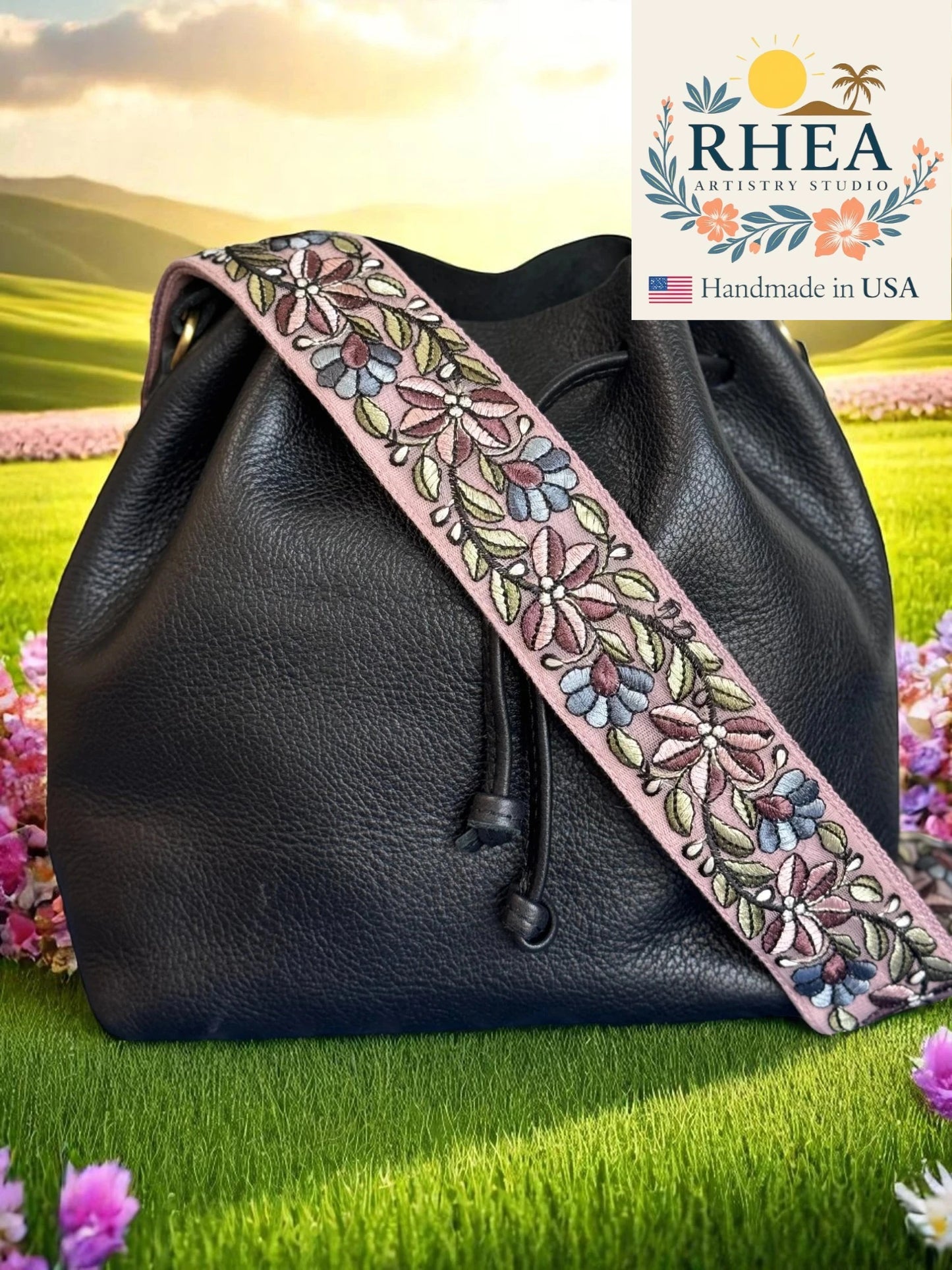 Black leather bag with a floral purse strap on grass with flowers and a scenic background, featuring the Rhea Artistry Studios logo.