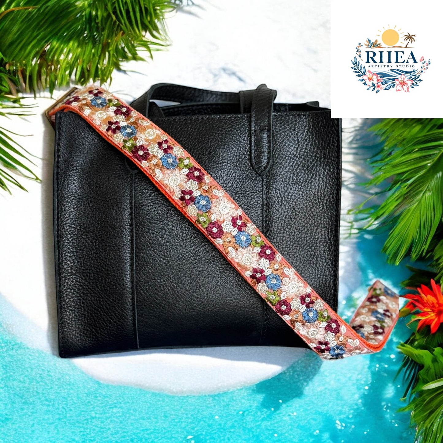 Black leather handbag with a coral strap embroidered in burgundy, blue, ivory, and green floral designs, set against a tropical sandy beach backdrop.