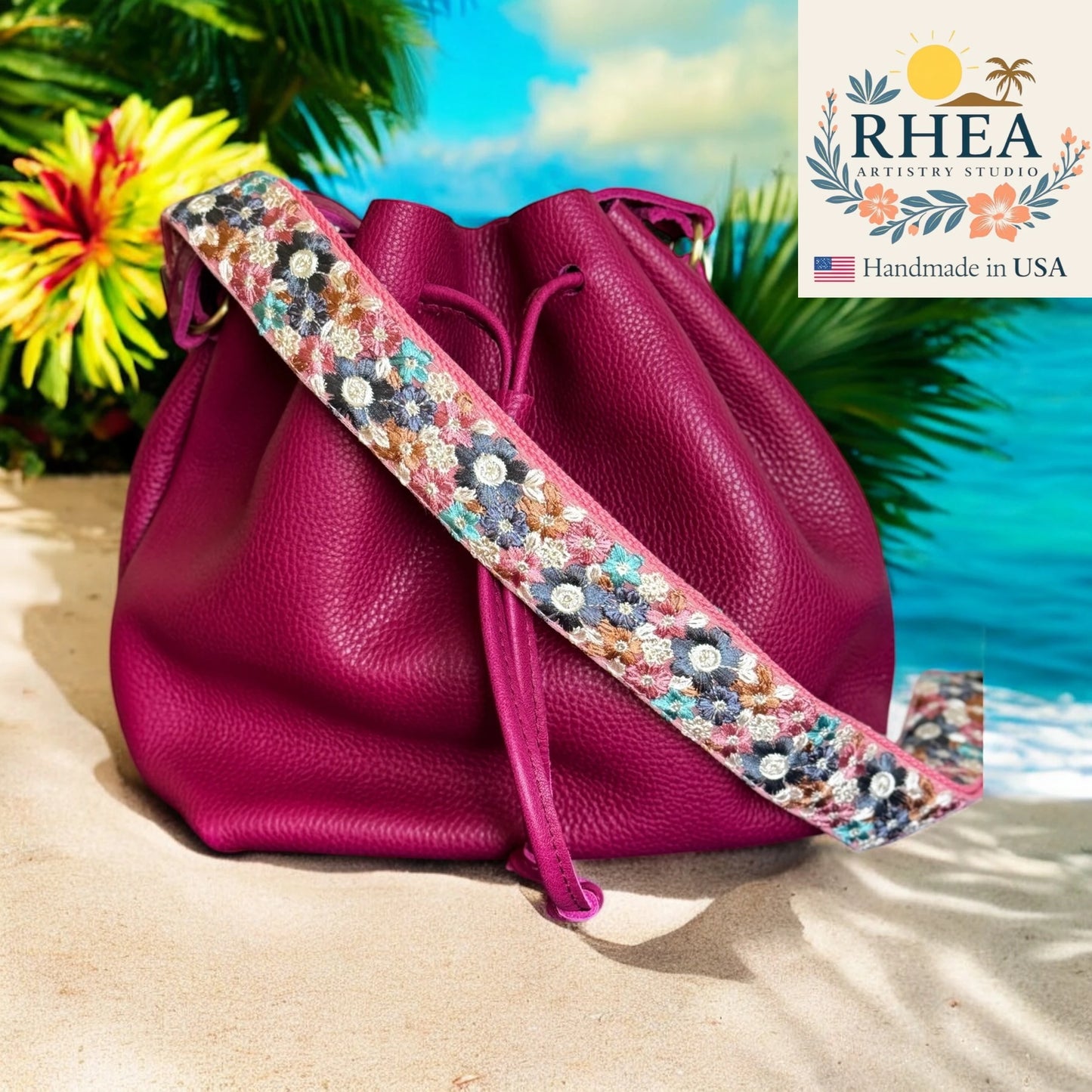 Pink leather bag with a floral purse strap on a beach, featuring the RHEA Artistry Studio logo.