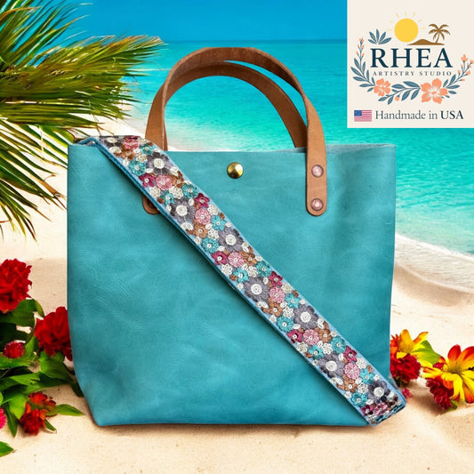 Blue tote bag with floral purse strap on a beach background, featuring the RHEA Artistry Studio logo.
