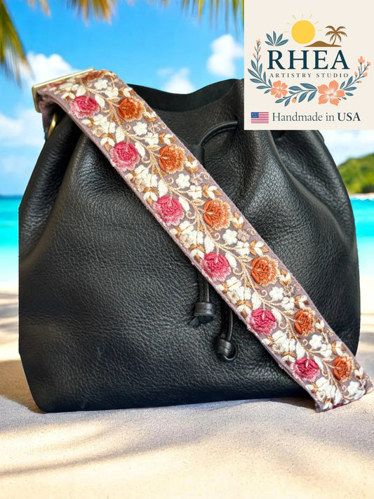 Black leather bag with a floral purse strap on a beach, featuring the Rhea Artistry Studio logo.