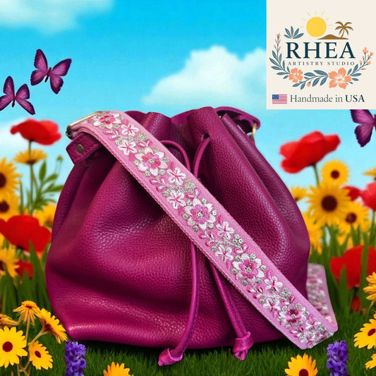 Purple leather bag with floral strap in a garden setting, featuring RHEA Artisan Studio logo.