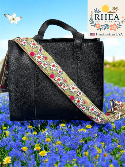 Black handbag with a floral purse strap in a field of flowers, featuring RHEA Artisan Studio branding.