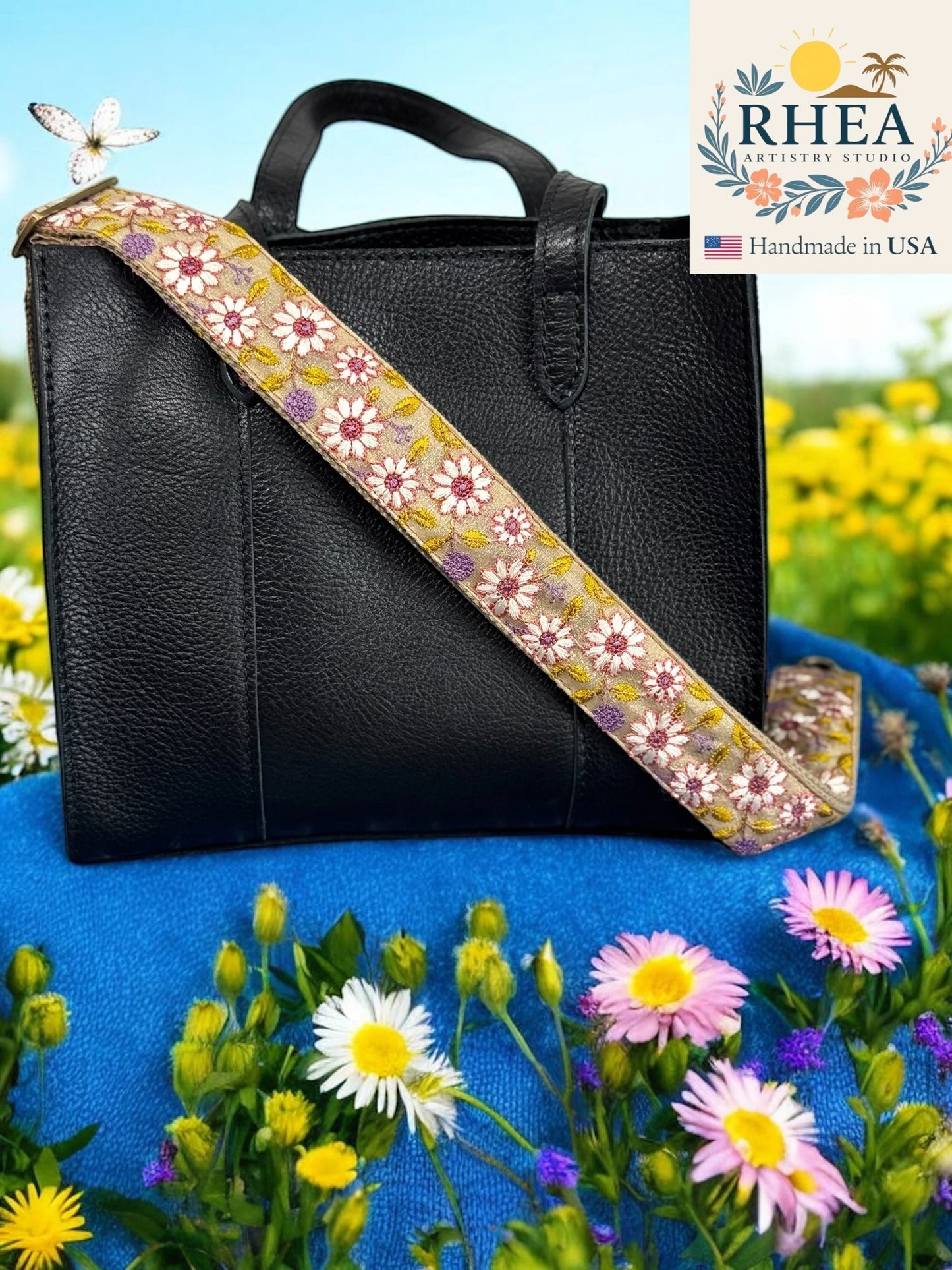 Black handbag with a floral purse strap on a blue surface with flowers, featuring the RHEA Artistry Studio logo.