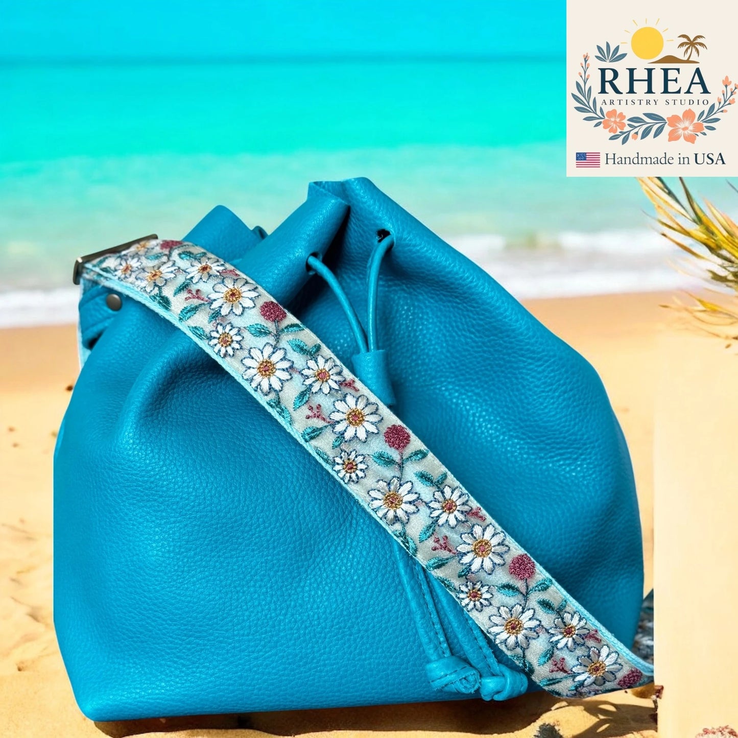 Blue leather bag with floral purse strap on a beach, featuring RHEA brand logo.