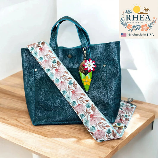 Teal handbag with floral strap and leather bag charm on a wooden surface, RHEA brand visible