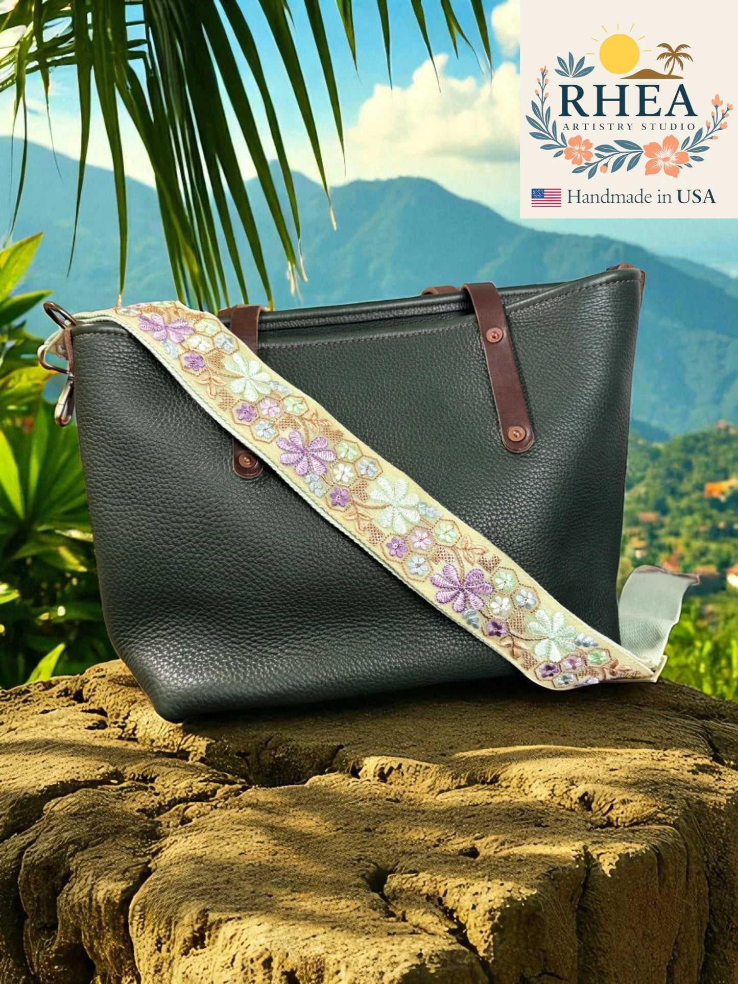 Black leather handbag with a floral strap on a rock with a scenic background, featuring the RHEEA brand.