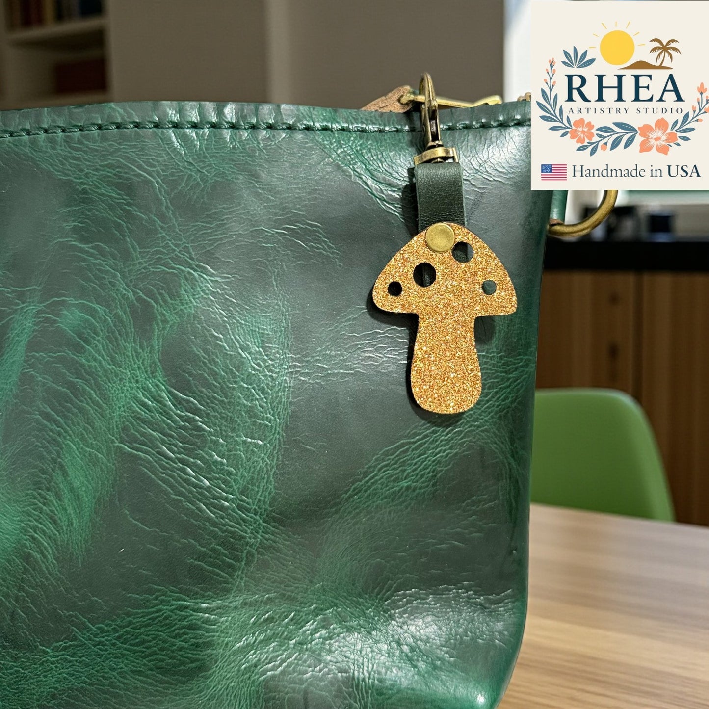 Green leather bag with a gold glittery mushroom keychain charm on a wooden surface, RHEA brand visible.