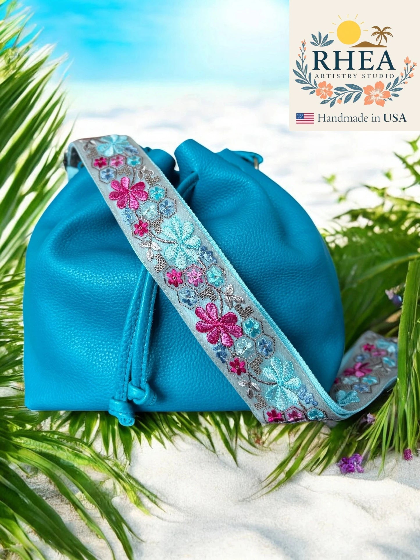 Blue bag with floral embroidery on a leafy background, featuring the RHEA brand.